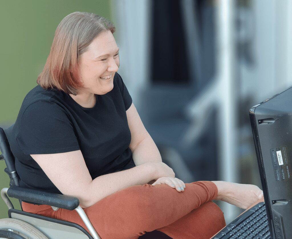 Picture of a woman in a wheelchair laughing , with a black t shirt and brown trousers.